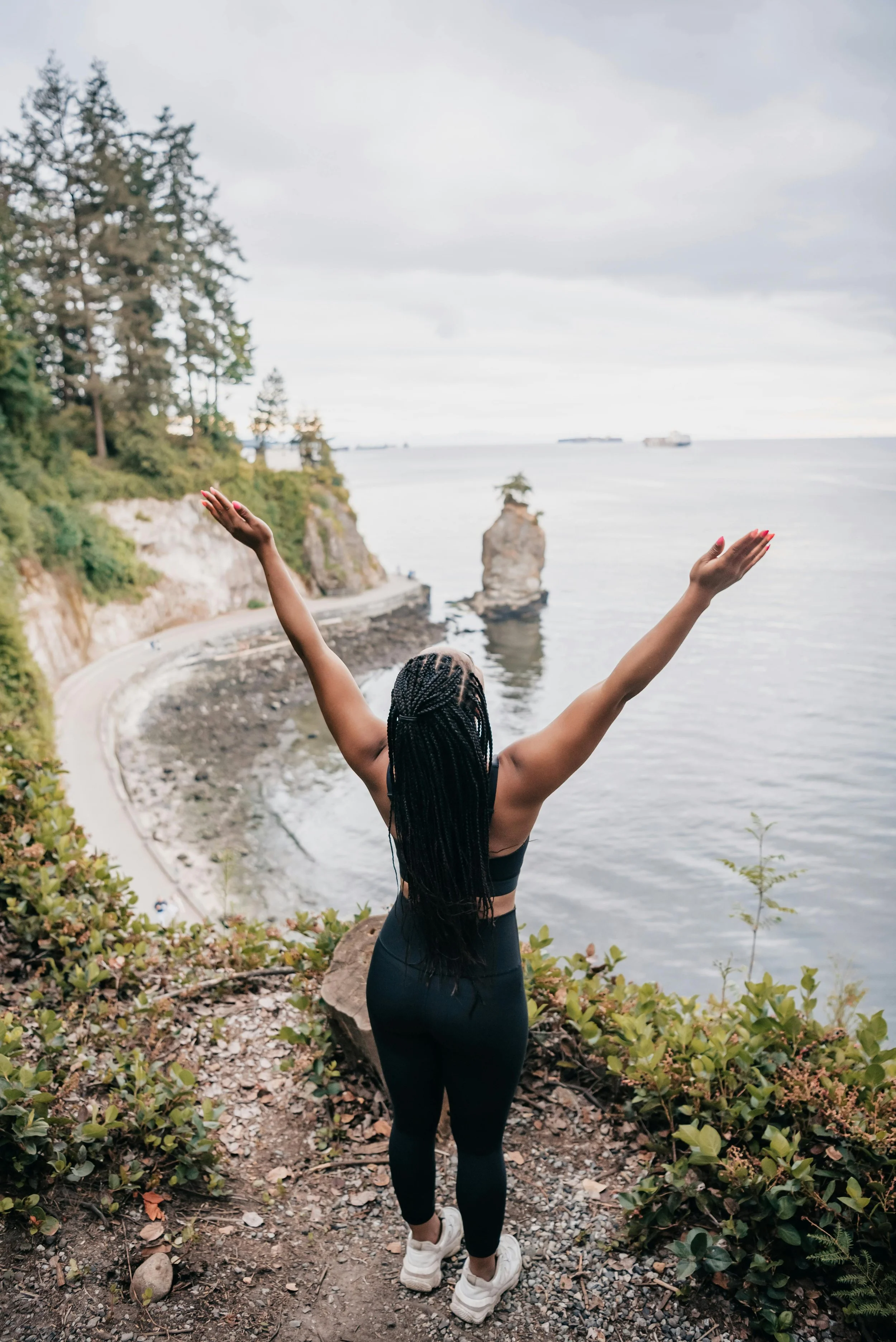 A woman standing on a cliff overlooking a coastal scene with trees, rocks in the water, and ships in the distance, with her arms raised in celebration.