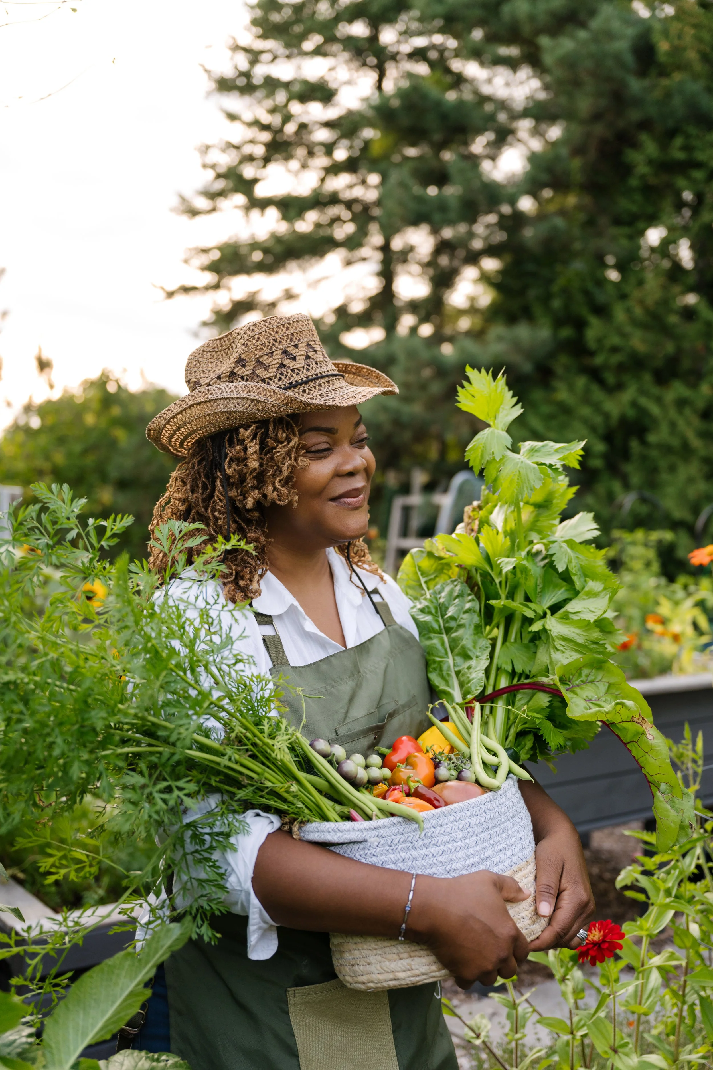 A woman with curly hair wearing a straw hat, white shirt, and green apron, holding a large basket of fresh vegetables in a garden.