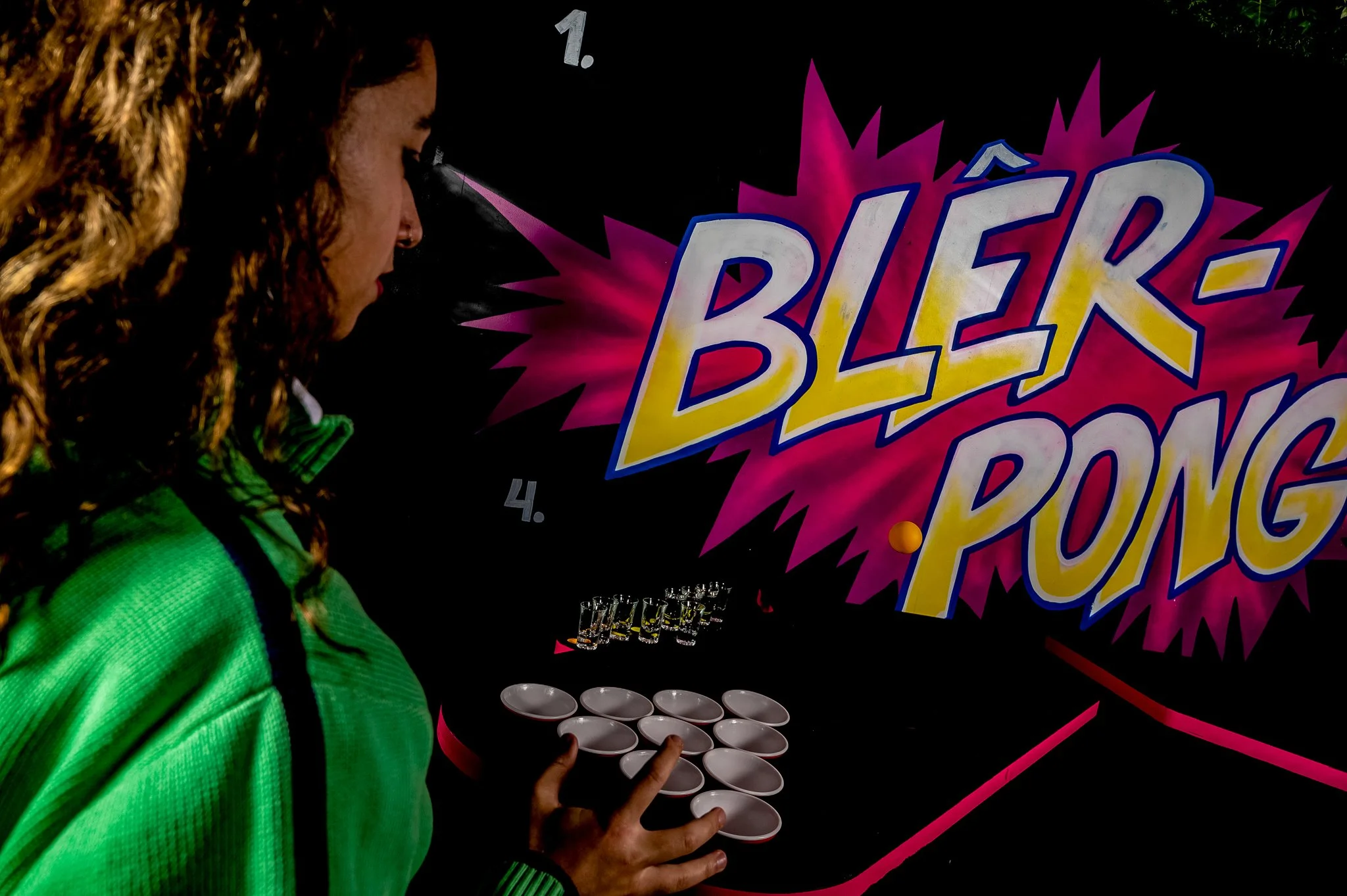 A young woman in a green jacket and curly hair playing a game of beer pong, with a colorful comic-style sign that reads 'BLER PONG' in the background.