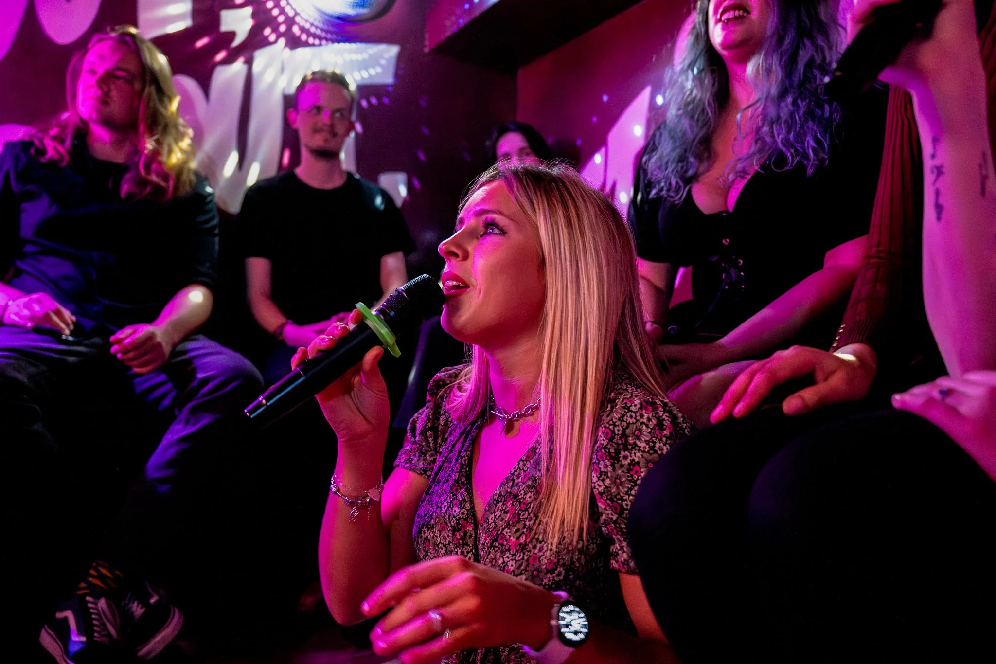 Young woman singing into a microphone during a karaoke session or live music event, surrounded by cheering friends in a dimly lit room with vibrant stage lighting.