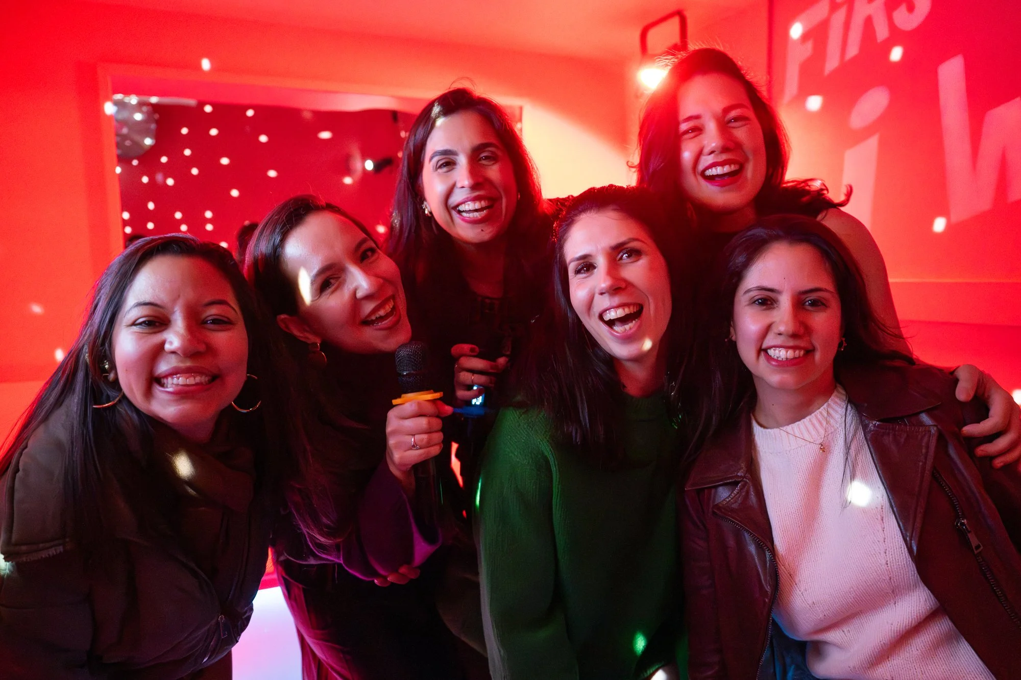 Group of seven women smiling and posing together at a party or celebration, with red lighting and a festive background.
