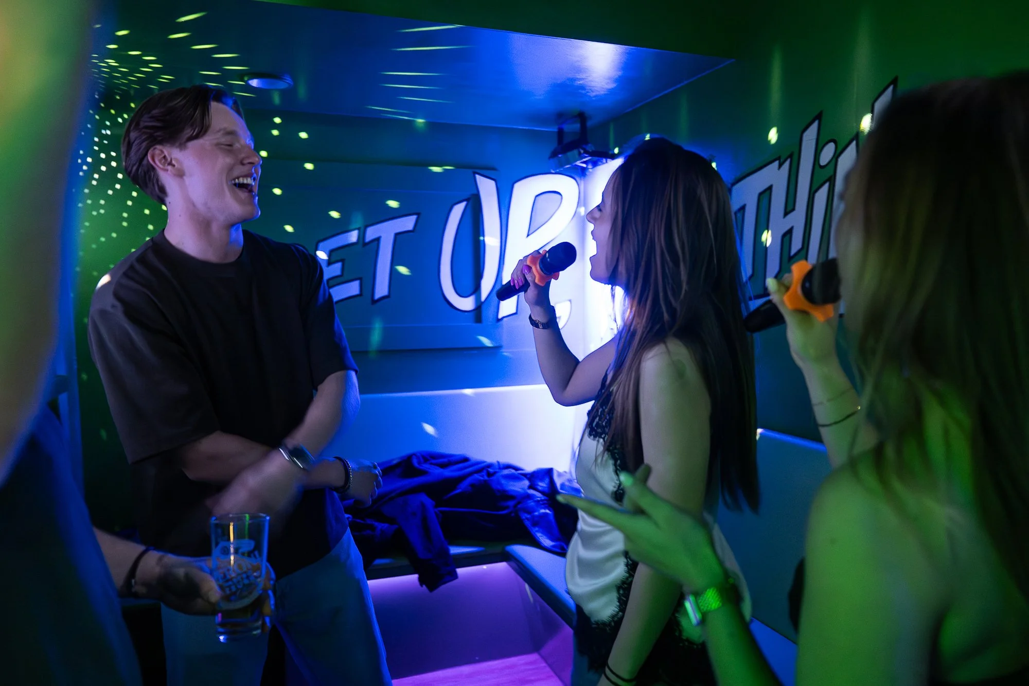 Four young adults at a karaoke bar, two women holding microphones singing and laughing, a man with a drink smiling, and another person partially visible holding a phone. Neon lights and a sign with the text 'GET UP' are in the background.