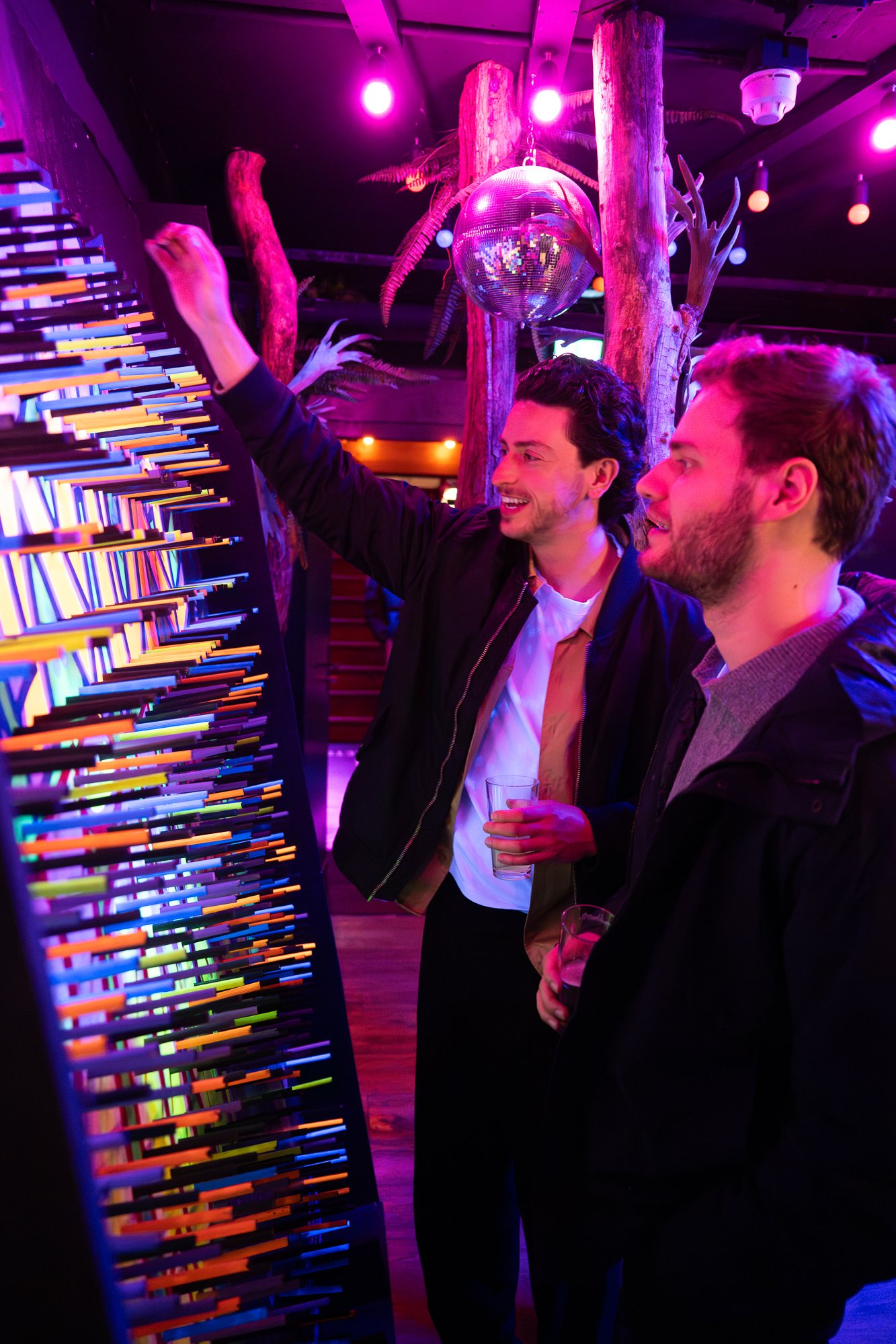 Two men stand in a colorful, neon-lit room lookingOne man is smiling and reaching towards the display, while the other holds a drink. The background has a disco ball hanging from the ceiling and wooden logs with plants, creating a vibrant, modern atmosphere.