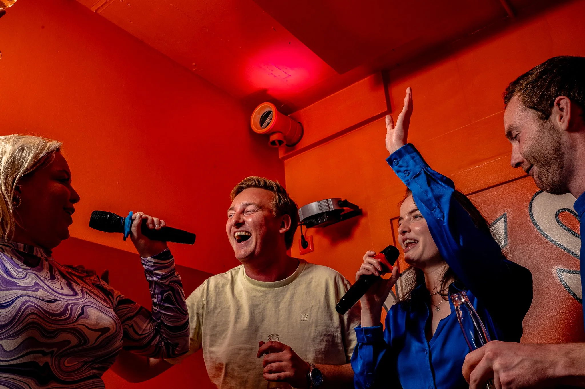 Four people singing and celebrating at a karaoke bar with orange walls, holding microphones and drinks, one woman raising her hand.
