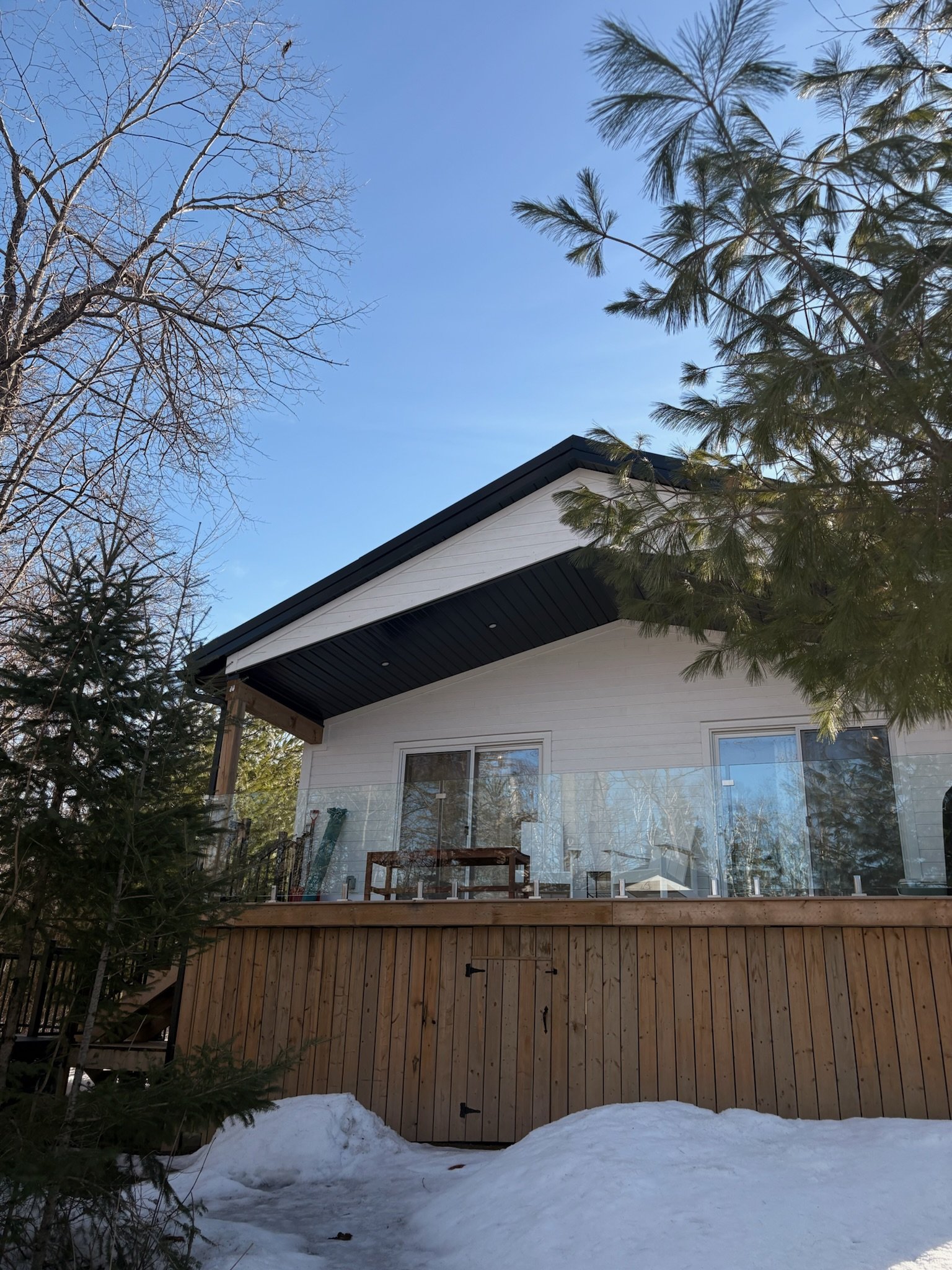 A house with a wooden deck and glass railing, surrounded by snow and trees, under a clear blue sky.
