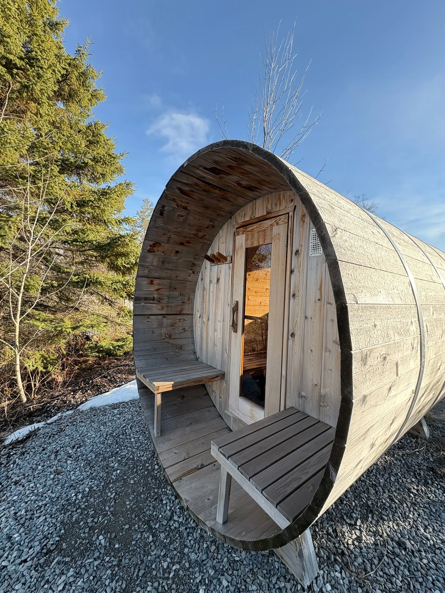 A wooden barrel-shaped cabin with a glass door, small benches on the exterior, surrounded by trees and gravel under a blue sky.