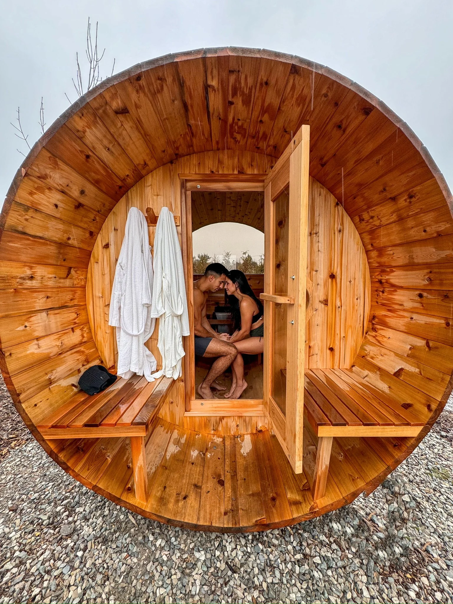 Two people sitting inside a small, round, wooden sauna, touching foreheads, with white robes hanging outside and a small black bag on the bench outside.