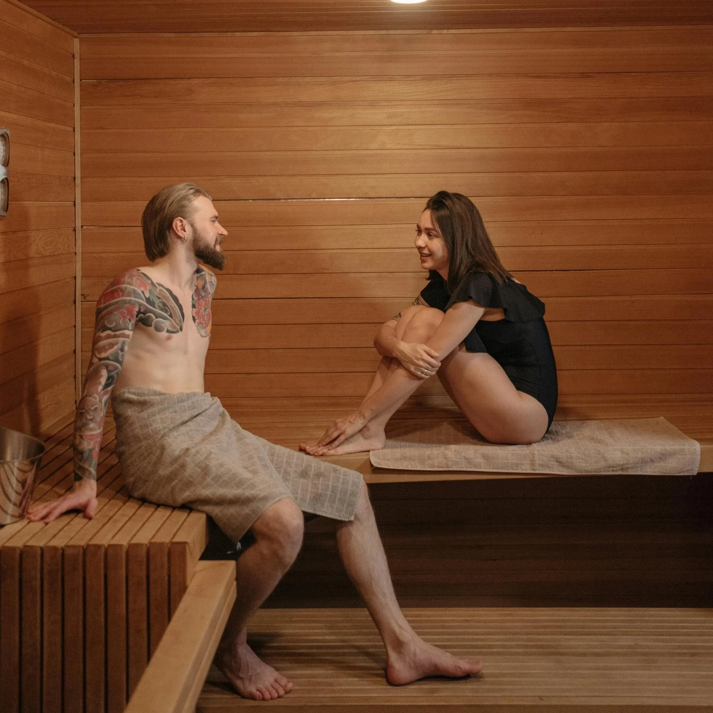 A man and woman sit inside a wooden sauna, smiling at each other. The man has long hair, a beard, and tattoos on his arms and chest, and is wrapped in a towel. The woman has shoulder-length brown hair and is wearing a black outfit, sitting with her knees drawn up and hands resting on her legs.