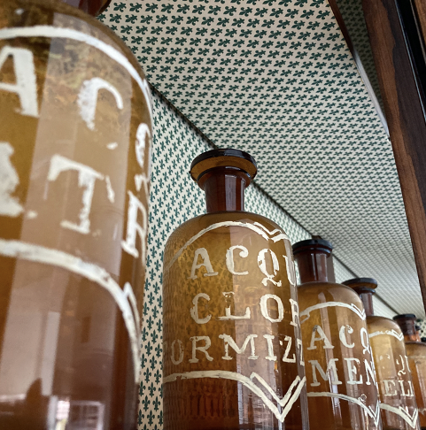 Serie di bottiglie di vetro marroni con etichette che leggono 'ACCLIMATEMENT' in un ambiente con soffitto decorato con un motivo a quadri verdi e bianchi.