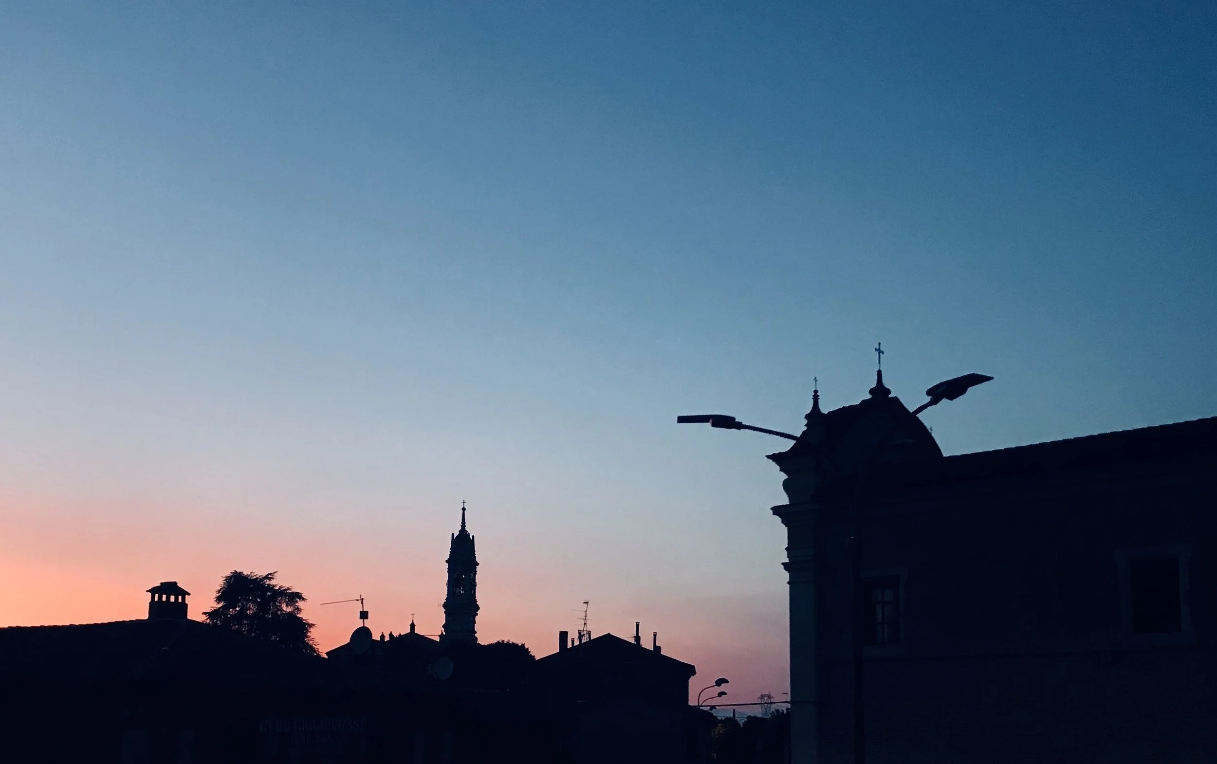 Sfumato al tramonto sopra tetti di un centro cittadino, con silhouette di chiese e muri, cielo blu e sfumature rosa abbagliata sullo sfondo.