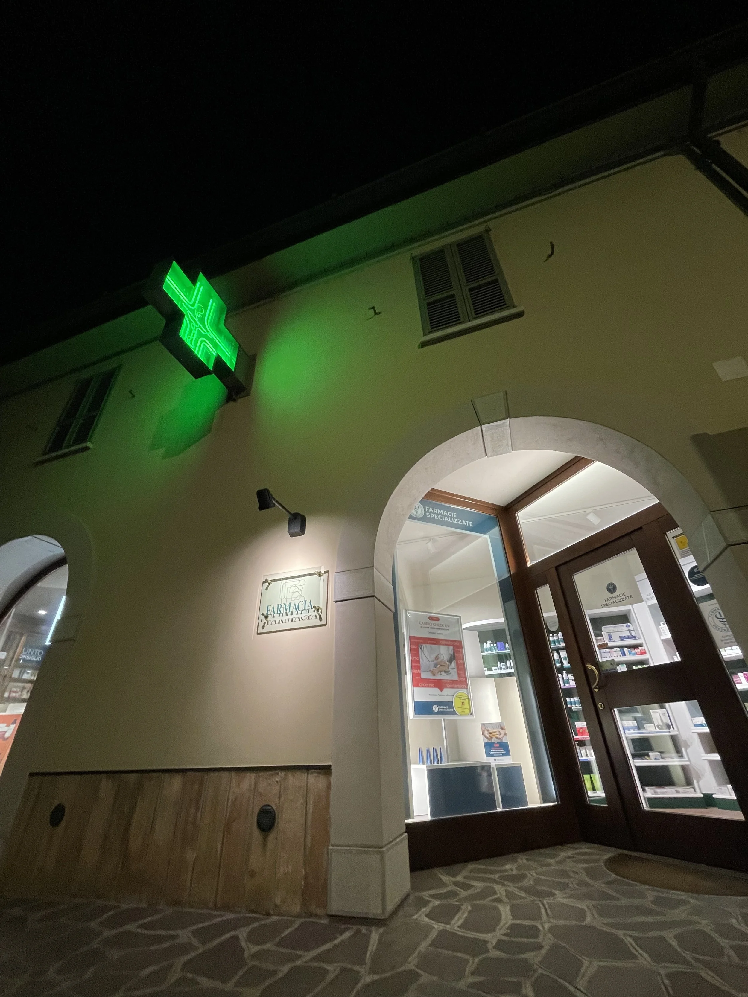 Esterno di una farmacia con insegna a croce verde illuminata e ingresso con vetrina e porta di vetro, di sera.