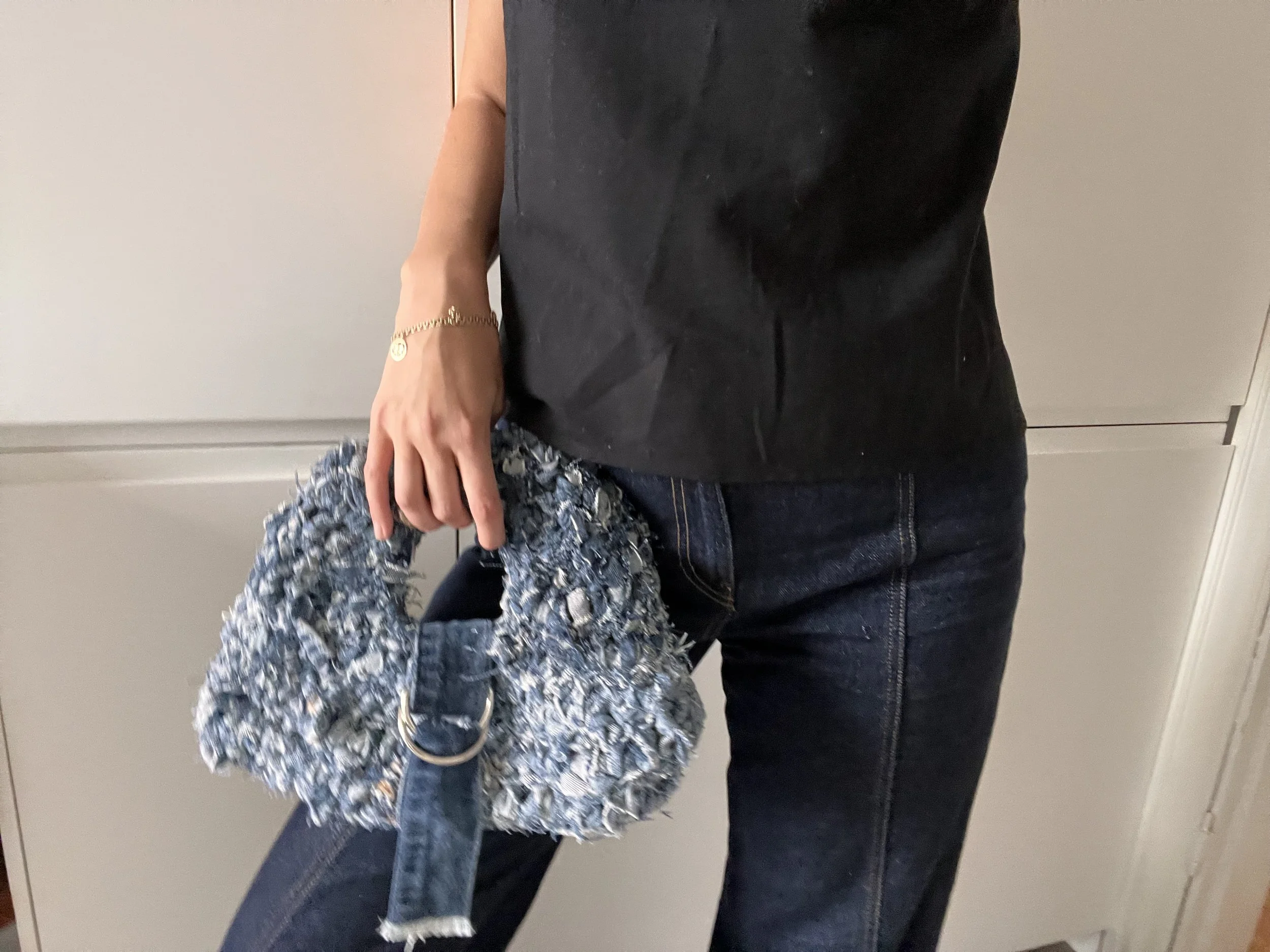 Person standing indoors holding a textured handbag with a shoulder strap, dressed in dark jeans and a black top.