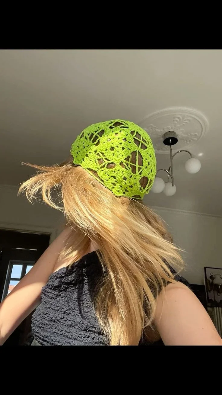 A person wearing a bright green crocheted hat, with long blonde hair flowing, indoors with a ceiling light fixture in the background.