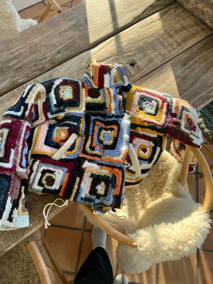 Colorful, multi-patterned crocheted blanket with square designs draped over a wooden table and a cream, fluffy chair.