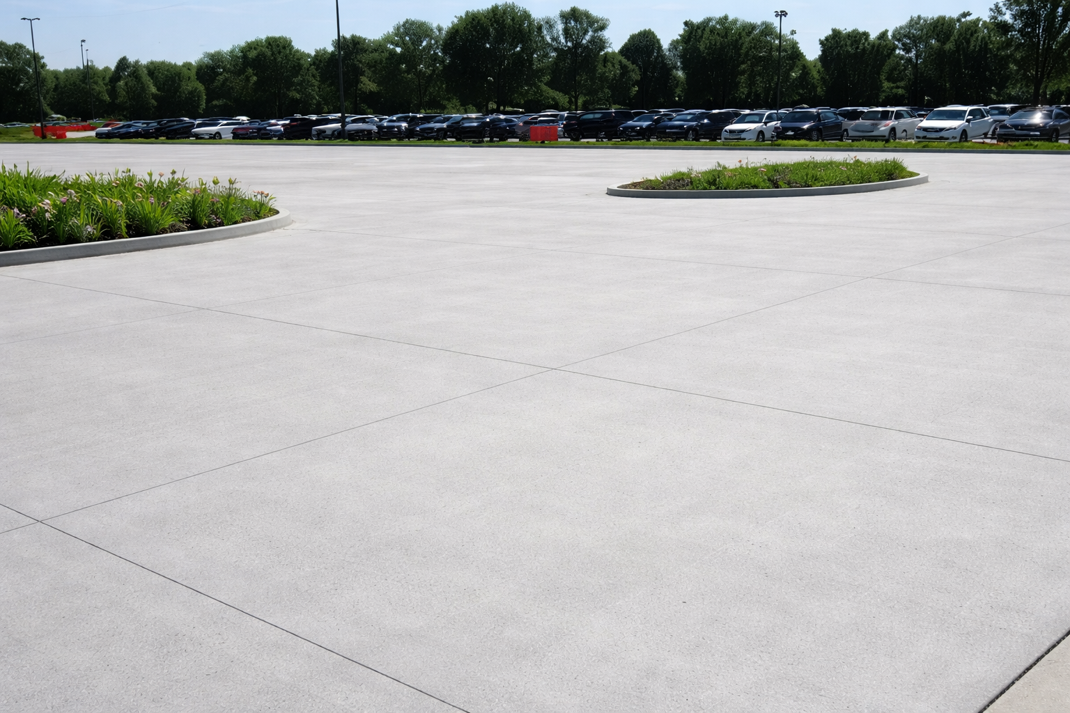 A parking lot with a large, empty concrete area, surrounded by a few small landscaped islands with flowers. In the background, there are many parked cars and trees, under a clear sky.