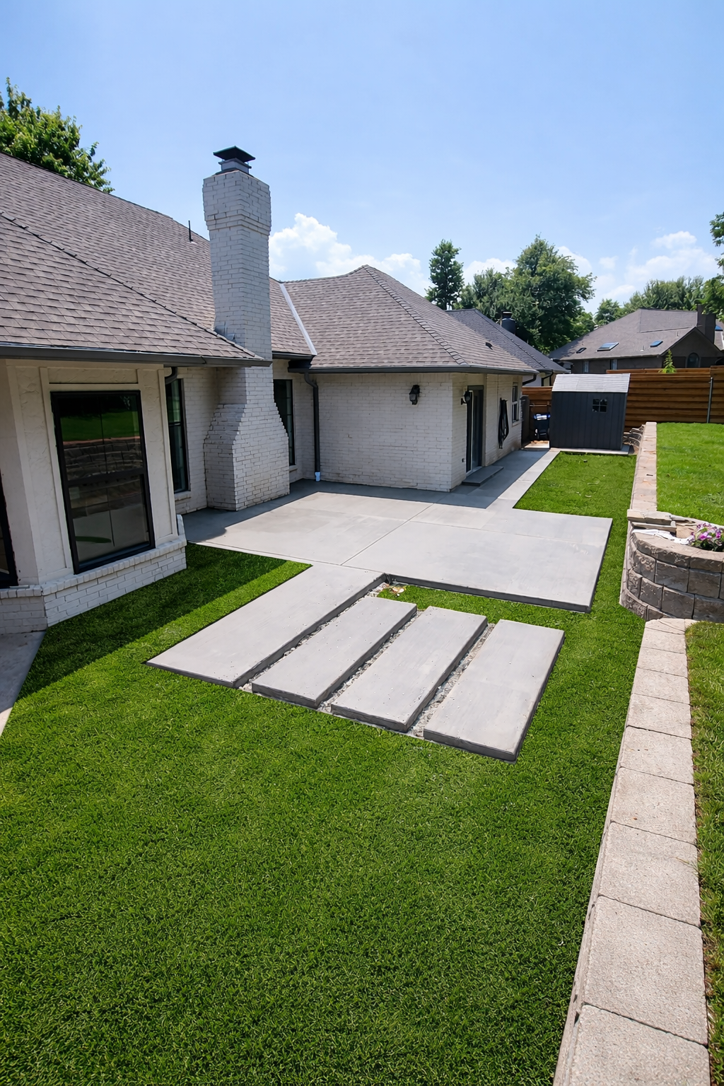 Backyard patio area with newly poured concrete, temporary concrete forms, green grass, a brick firepit, a chimney, a small shed, and a wooden fence under a clear blue sky.