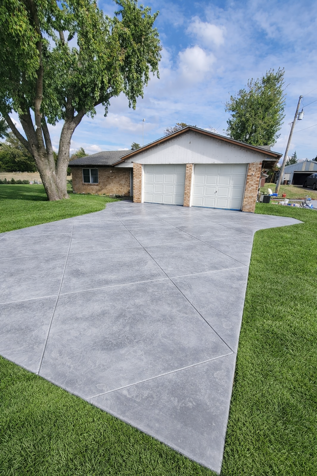 Concrete driveway leading up to a brick garage with two doors, surrounded by green grass and trees under a partly cloudy sky.