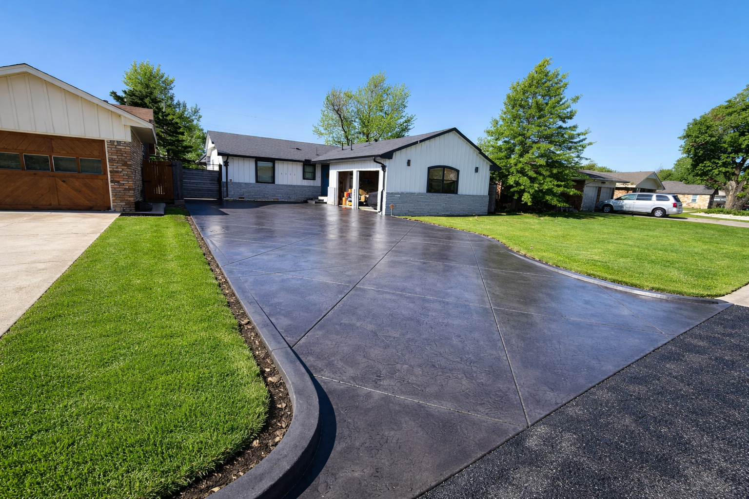 Front yard of a house with a freshly paved driveway, green lawns, and trees under a blue sky.