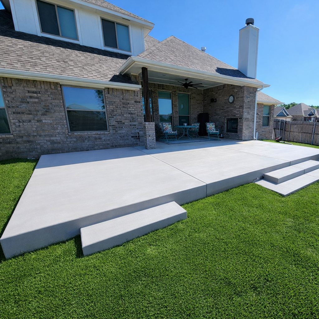Backyard with concrete patio steps leading up to a covered patio area of a brick house, with outdoor furniture and a ceiling fan, under a blue sky.