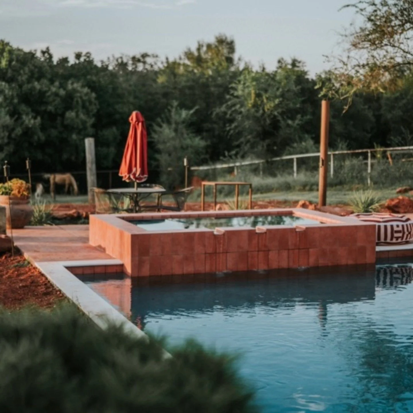 A backyard with a small pool and a hot tub, surrounded by a brick patio, with a red umbrella and outdoor furniture, and trees in the background.
