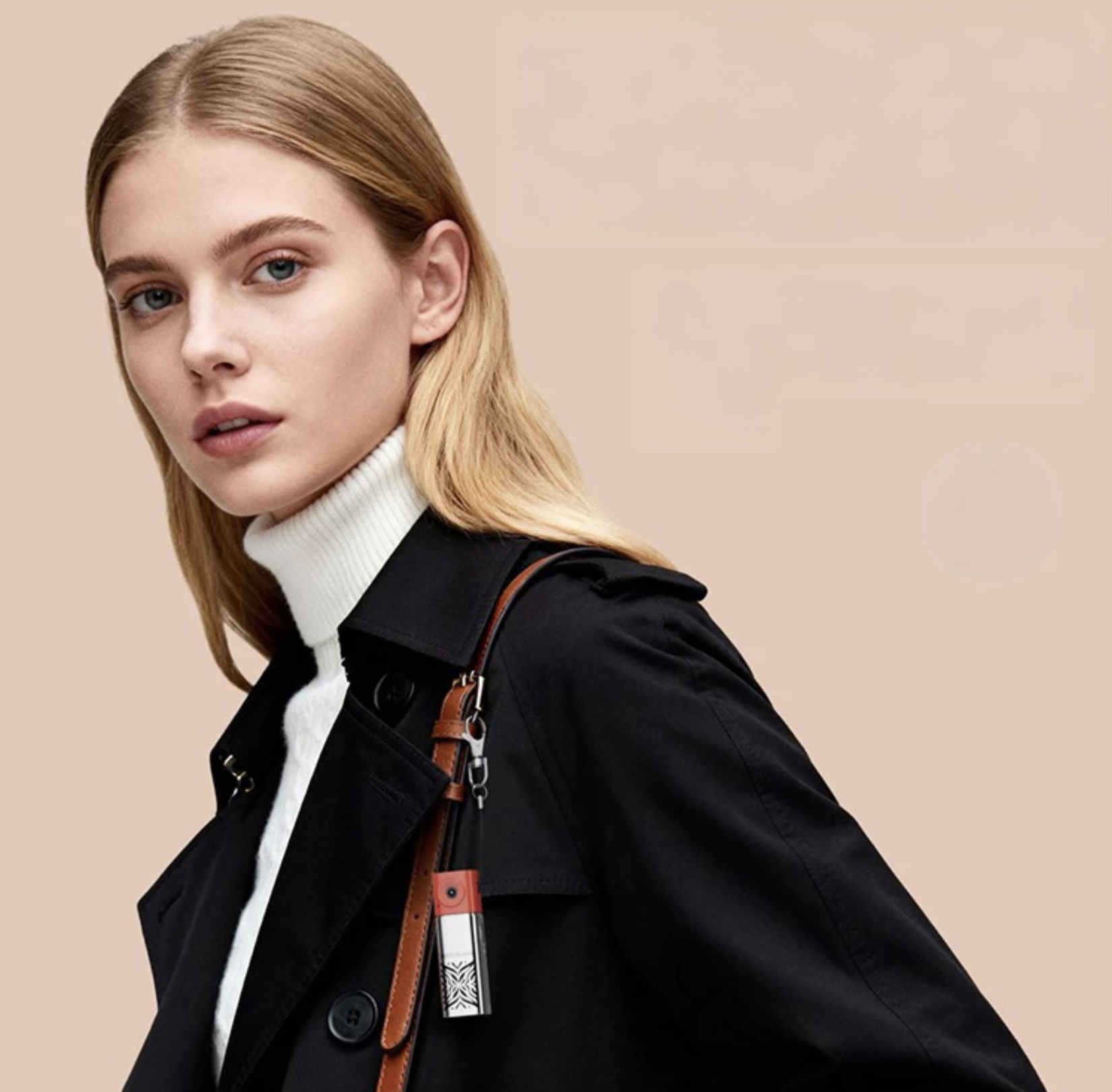 Femme avec cheveux blonds portant un manteau noir et un col roulé blanc, contre un fond beige.