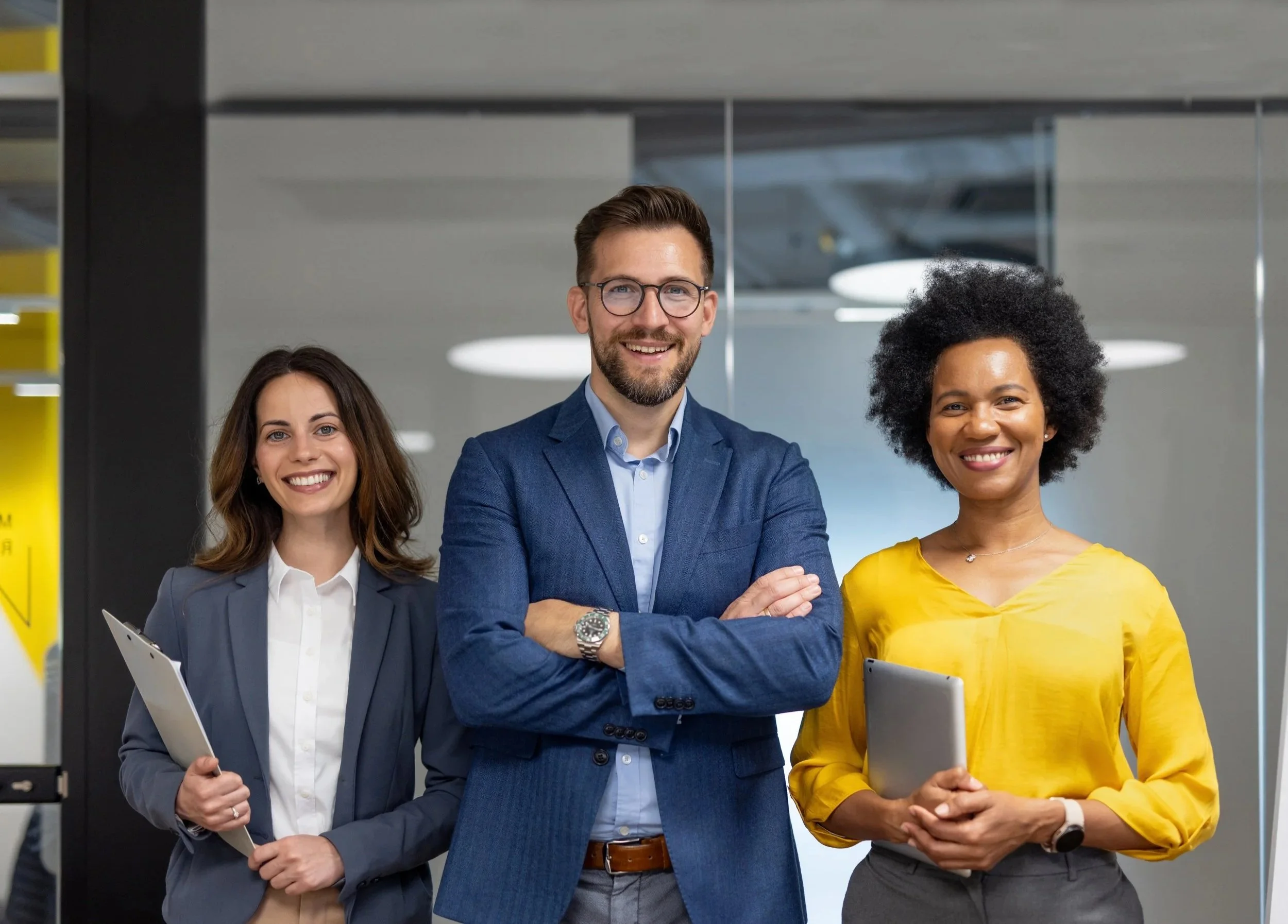 Three professionals posing confidently in an office setting. The man in the center has a beard, glasses, and is wearing a blue suit. The woman on the left has long brown hair and is wearing a grey blazer, holding a folder. The woman on the right has a large afro hairstyle, is wearing a yellow top, and a watch on her left wrist.