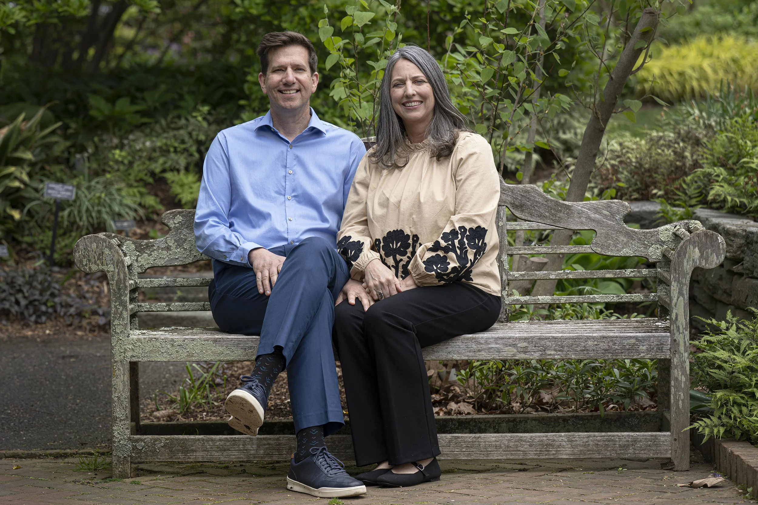 A smiling man and woman sitting on an aged wooden park bench in a garden surrounded by trees and bushes.