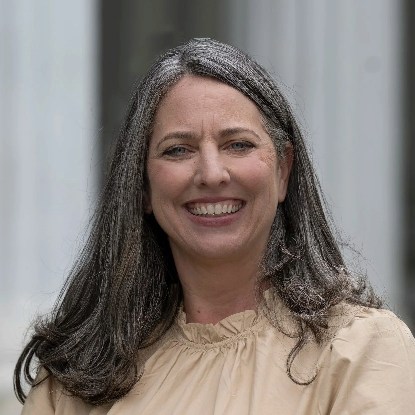 Close-up of a smiling woman with long wavy gray hair wearing a beige blouse.
