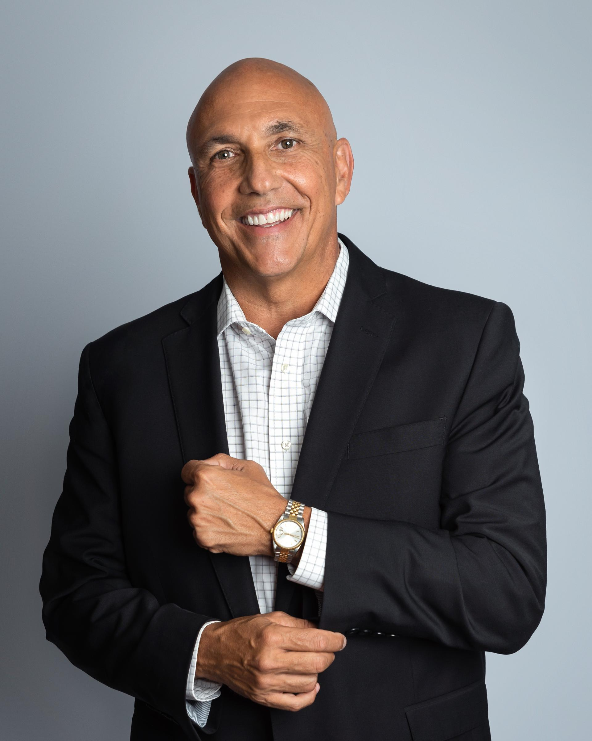 A smiling man in a black suit, white checkered shirt, wearing a gold watch, standing against a plain grey background.