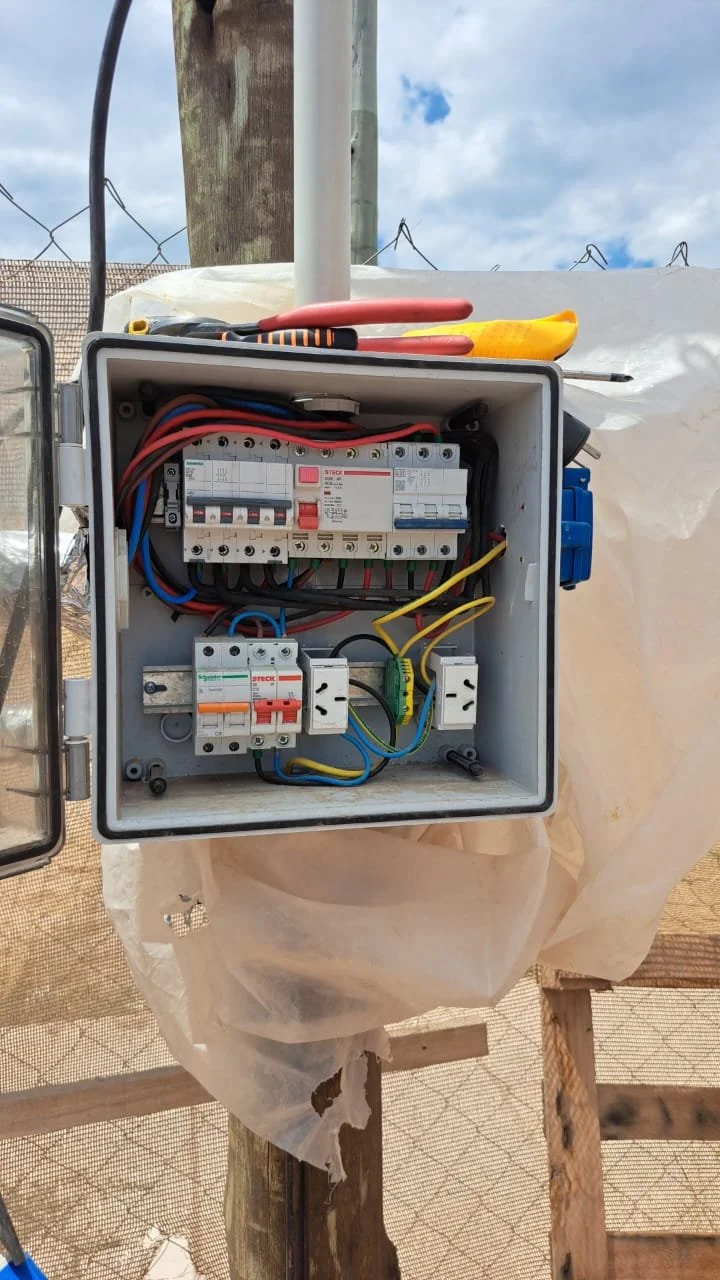 Caja de control eléctrico con componentes y cables, situada sobre una estructura de madera y cubierta parcialmente con plástico transparente.