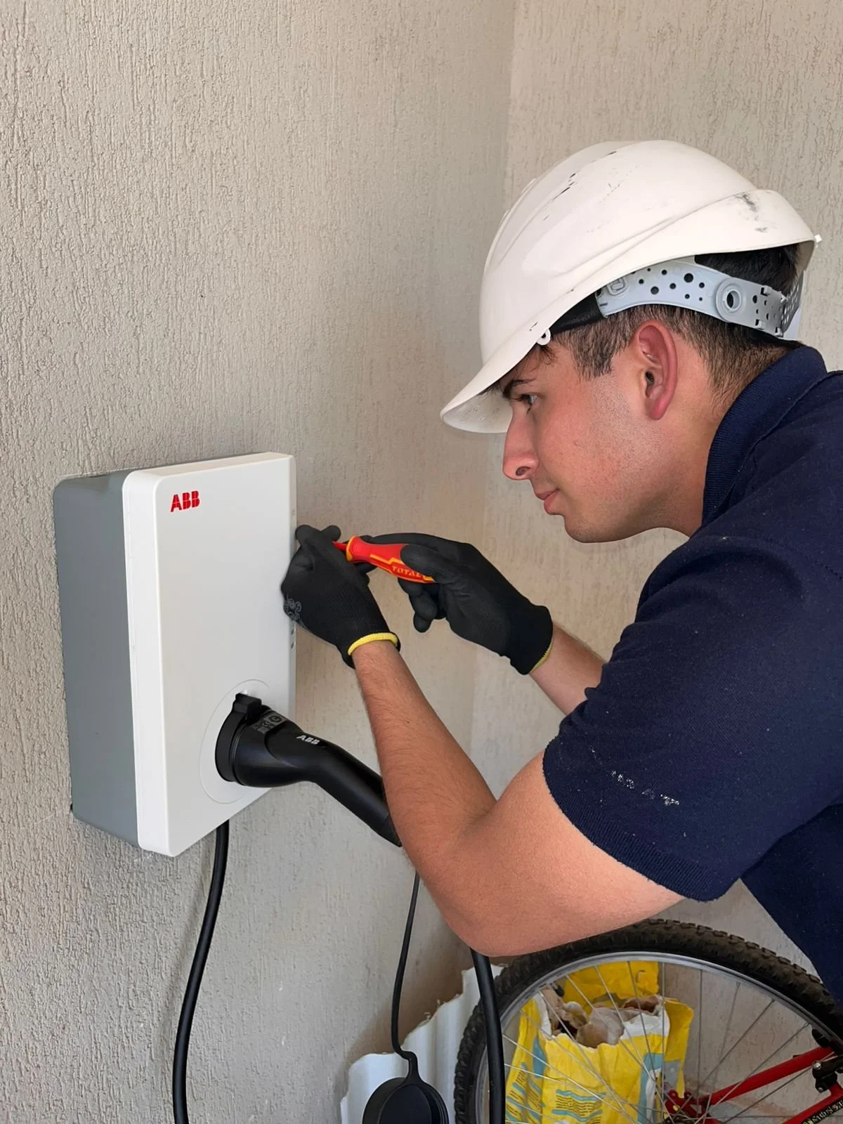 Un hombre usando casco y guantes trabajando en un cargador de coche eléctrico ABB con herramientas en mano.