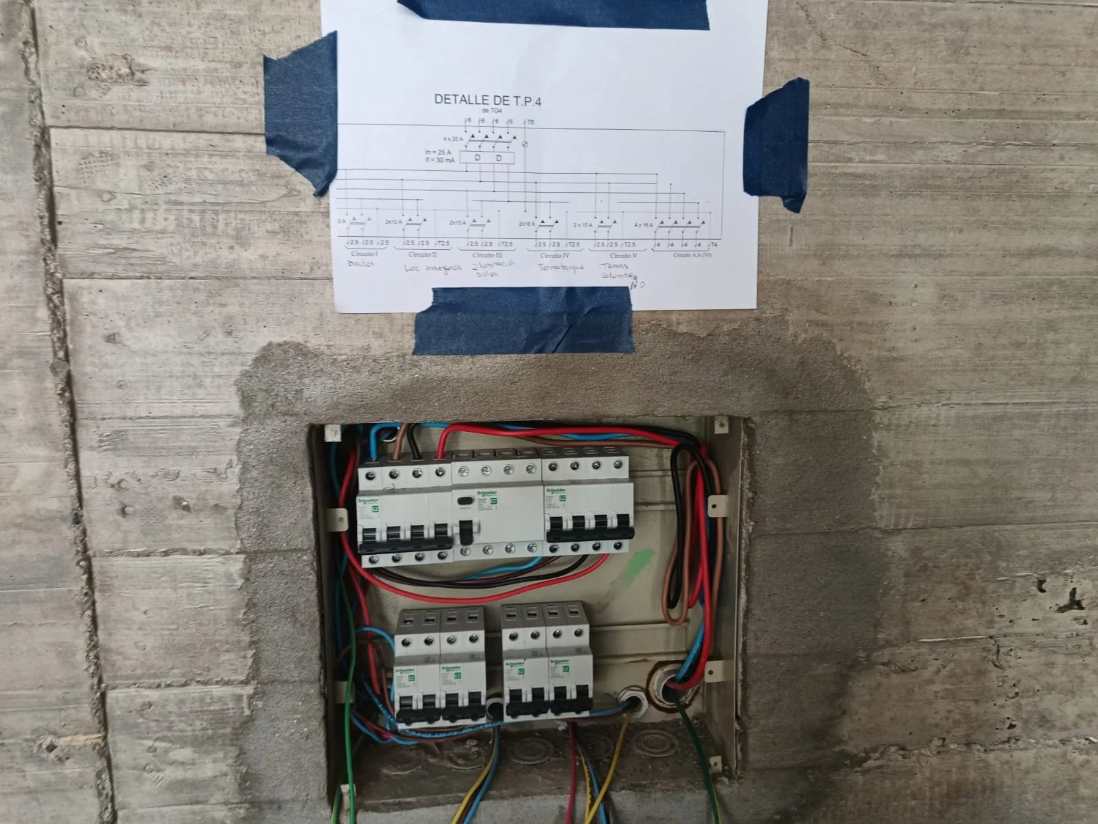 Tablero de distribución eléctrica con cables conectados y un esquema técnico en papel en la pared.