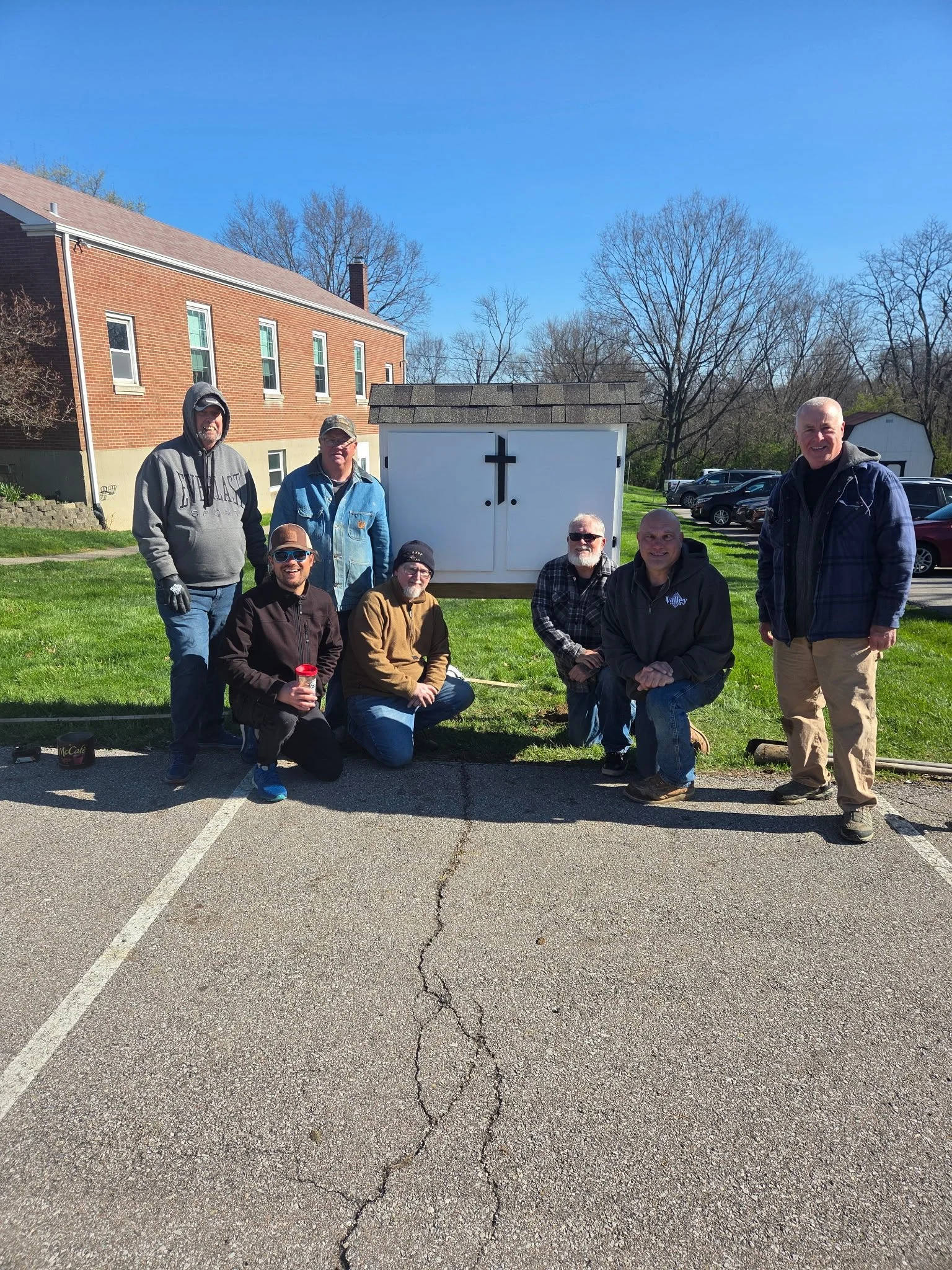 Group of seven men posing outdoors in front of a white cabinet with a cross, in a parking lot near a church or community building on a sunny day.