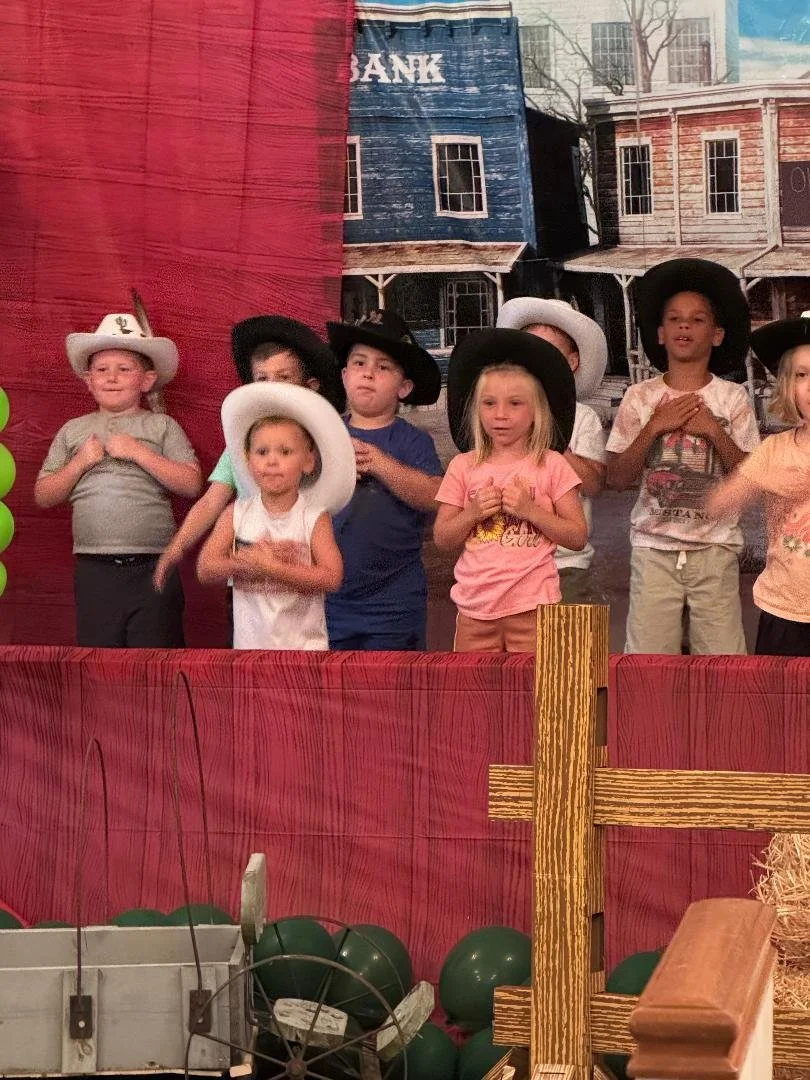 Children dressed in Western-themed costumes, wearing cowboy hats, standing behind a red curtain stage with a rustic Western town backdrop, likely performing a play or participating in a themed event.