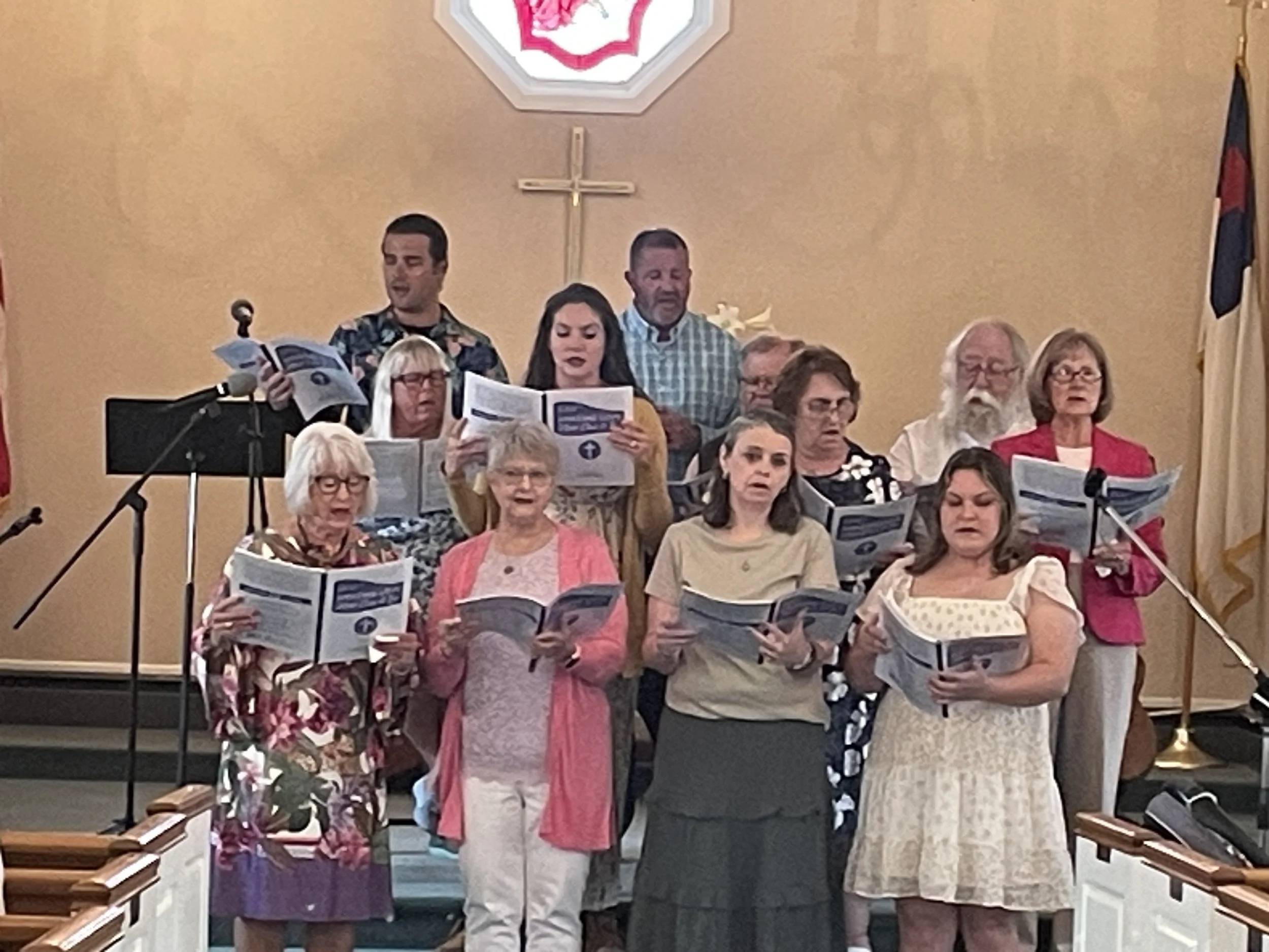 Group of people singing in a church choir, holding songbooks and standing on risers, with a cross on the wall behind them.