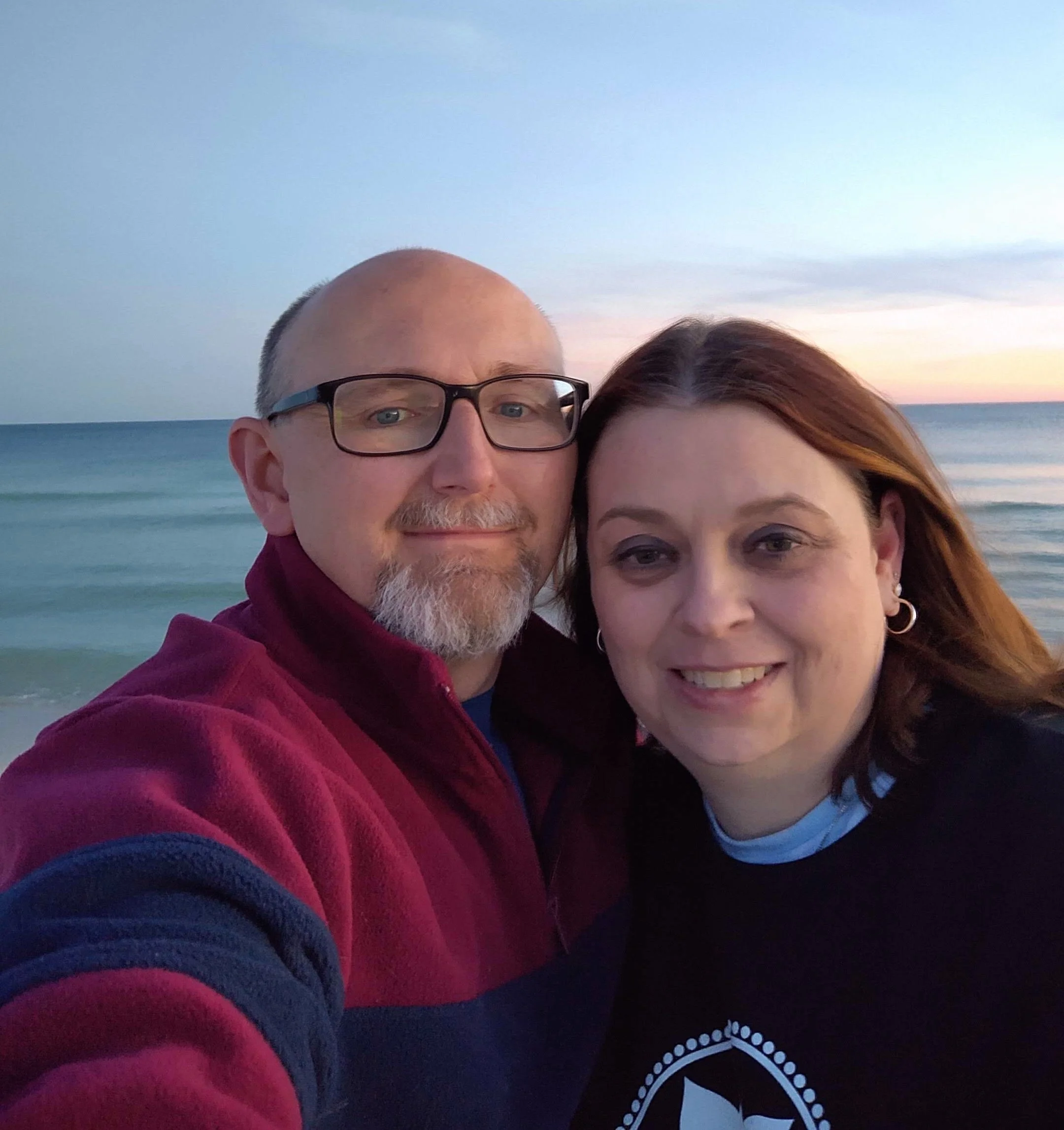 A couple taking a selfie on the beach at sunset. The man is wearing glasses and a red jacket, and the woman has auburn hair and is wearing a black shirt with a logo.