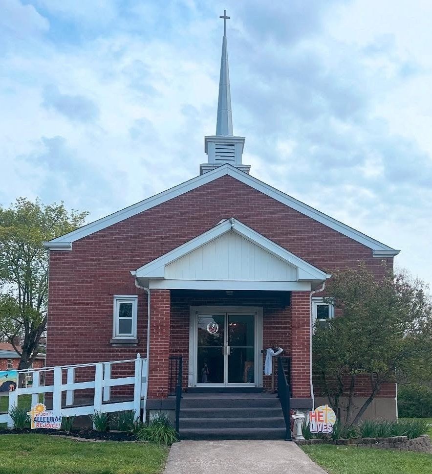 A small red brick church with a white steeple topped by a cross, white trim, and a front porch with steps and a glass door, surrounded by greenery and signs supporting religious celebrations.
