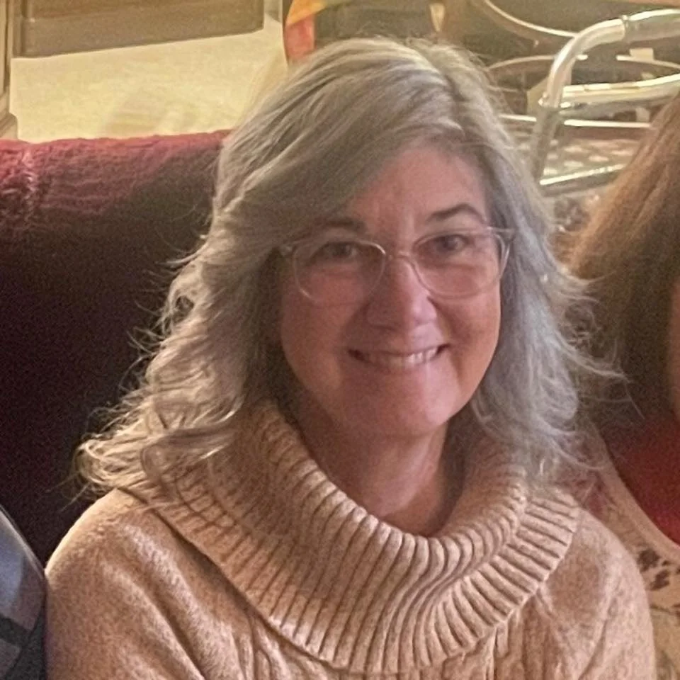 A woman with glasses and shoulder-length gray hair, smiling, wearing a beige turtleneck sweater, sitting on a red couch in a cozy room.