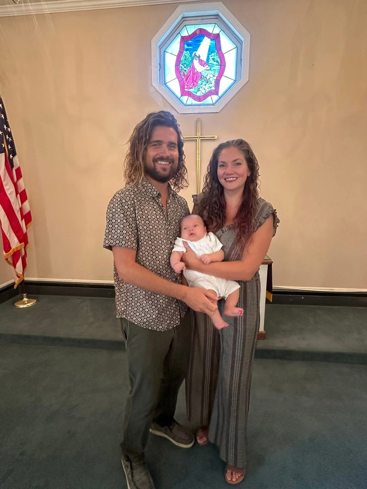 A family of three standing inside a church, holding a baby. The father has long, curly hair and a beard, wearing a patterned short-sleeve shirt. The mother has long, wavy hair, wearing a striped jumpsuit. The baby is dressed in white. Behind them is a stained glass window with a religious scene and a cross beneath it, with an American flag on the left side.