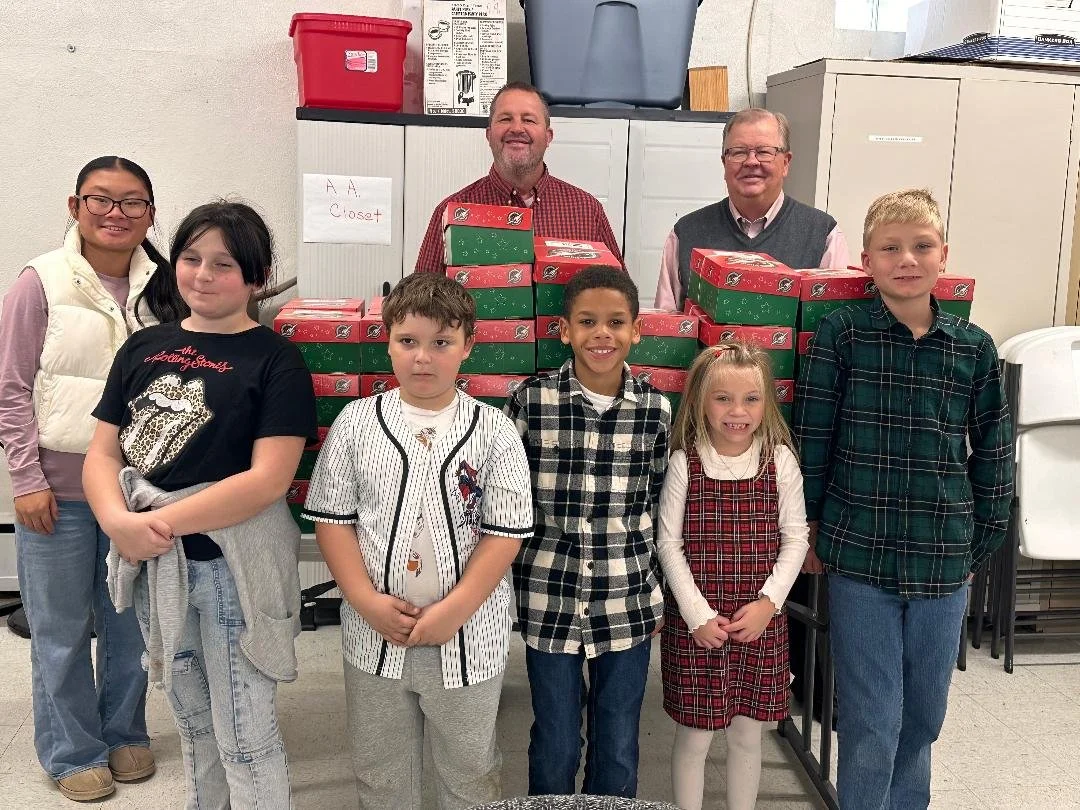 Group of seven children and two adults standing behind a table of pizza boxes, smiling in a room with storage cabinets.