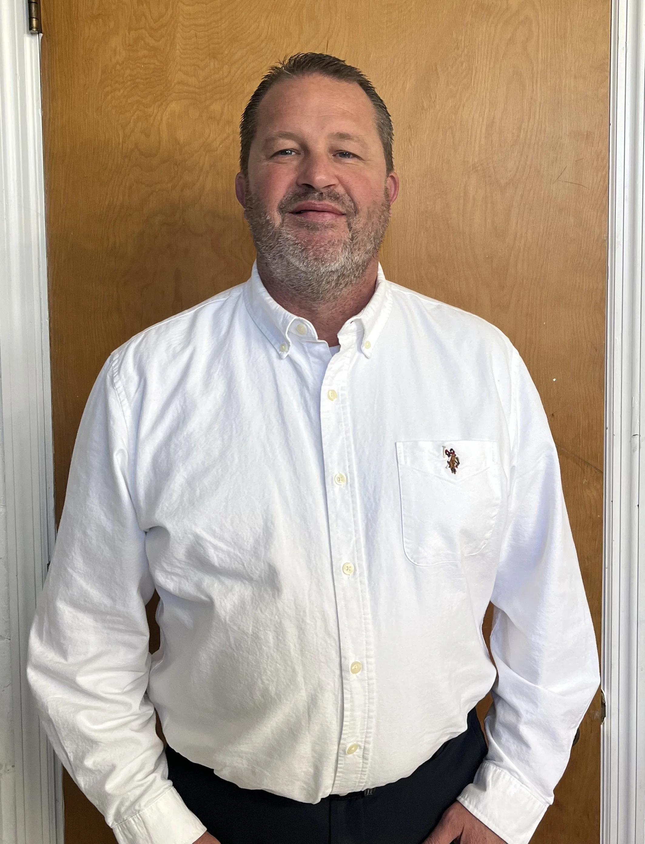A man with short dark hair and a beard, wearing a white button-up shirt with a small embroidered logo on the pocket, standing in front of a wooden door.