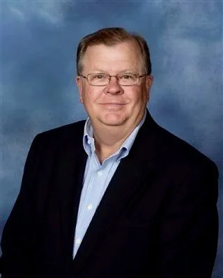 A middle-aged man wearing glasses, a dark blazer, and a light blue shirt, posing against a blue background.