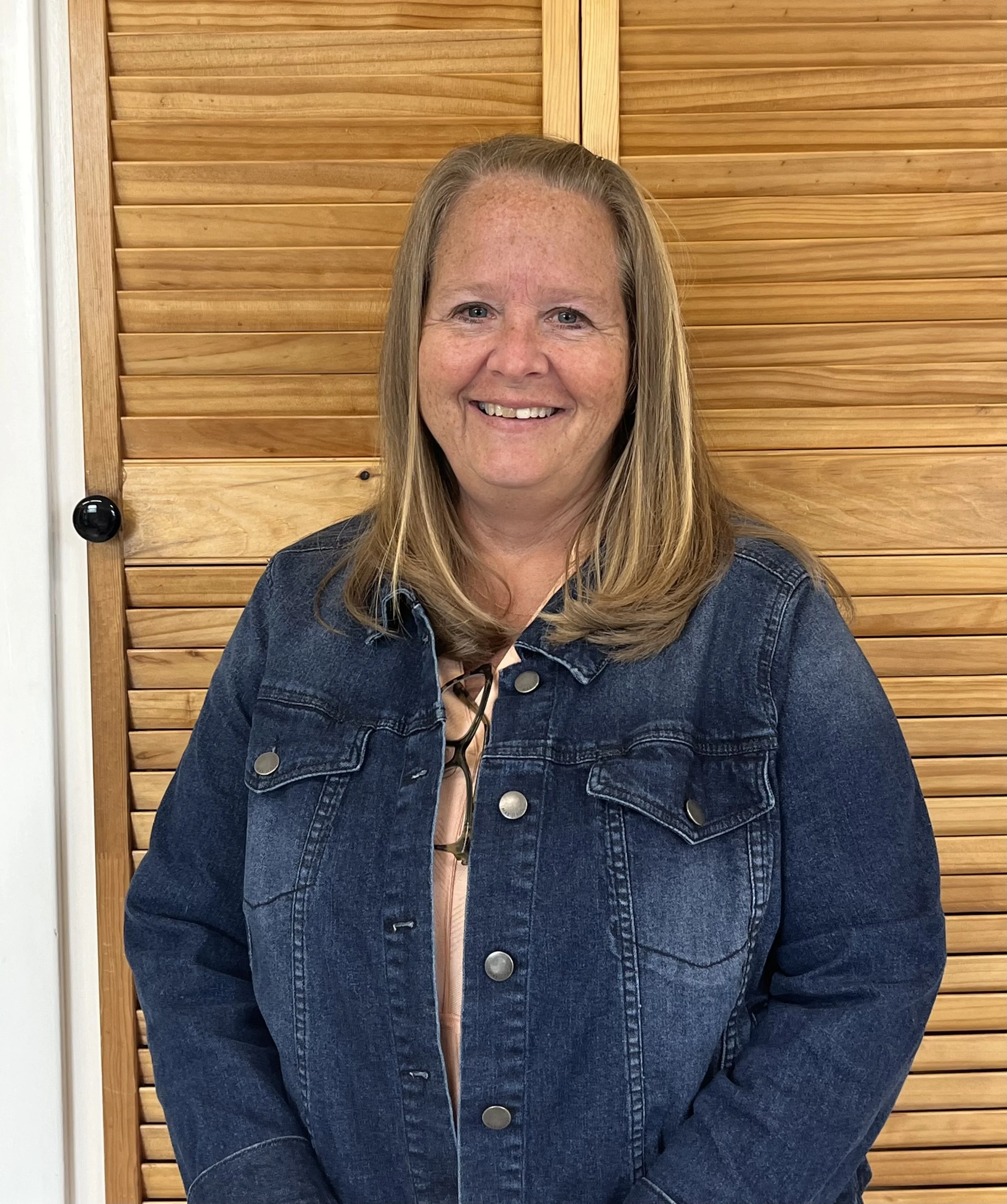 A woman with shoulder-length blonde hair smiling, wearing a denim jacket, standing in front of a wooden wall.
