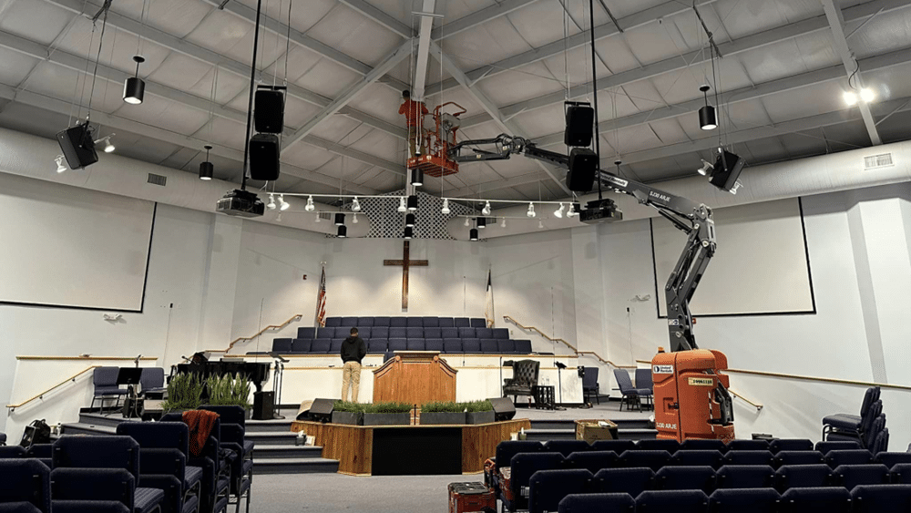 Interior of a church or auditorium with a stage, cross on the wall, seating, and equipment being set up, including a scissor lift.