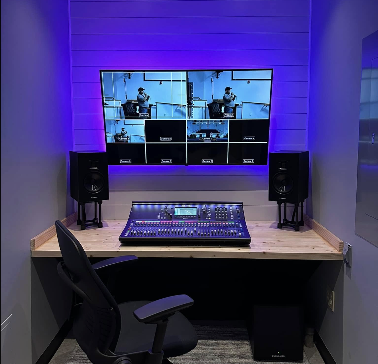 A control room with a mixing console on a wooden desk, two studio monitors on stands, a chair, and a large screen displaying a video feed with multiple camera views, illuminated by purple lighting.