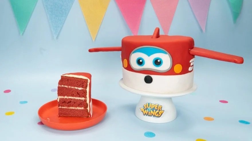 Ein Kuchen in Form eines Super Wings Flugzeugs, daneben ein Stück roten Kuchen, alles auf einer hellblauen Oberfläche mit bunten Papptellern und bunten Pompons, im Hintergrund bunte Fahnen.