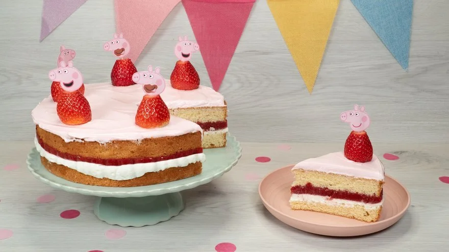 Geburtstagstorte mit Peppa Pig Figuren und Erdbeeren, daneben ein Stück auf einem kleinen Teller, bunte Fahnen im Hintergrund, pinke und rote Punkte auf dem Tisch.