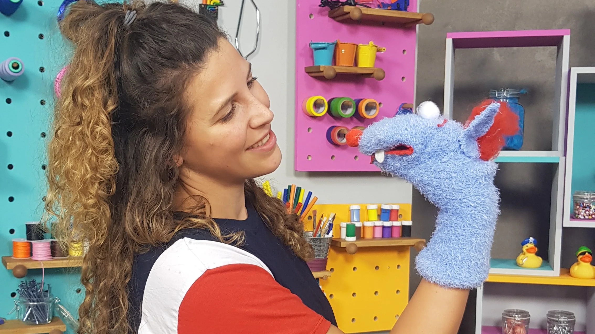 Eine junge Frau lacht, während sie mit einer Stoffpuppe spielt, in einem bunten Bastelraum mit verschiedenen Farben und Materialien.