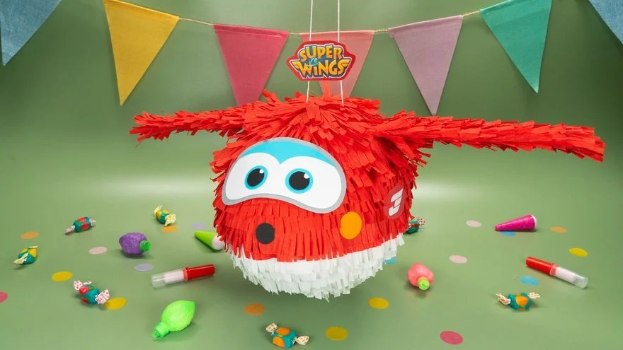 Bunter Piñata in Form eines Super Wings Charakters, umgeben von Partydekorationen wie Konfetti, Süßigkeiten, bunten Wimpeln und Partyhüten auf einem grünen Tisch.