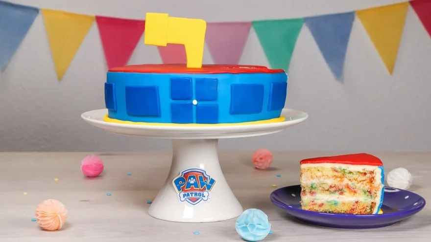 Ein bunter Geburtstagskuchen mit Paw Patrol Dekoration, daneben ein Stück Kuchen, dekorative Pappteller und sommerliche Dekorationen vor einer bunten Girlande.